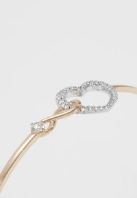 Metalinė apyrankė su auksinio atspalvio juosta ir begalybės simboliu, puošta širdies formos sidabriniu akcentu ir skaidriais krištolais.