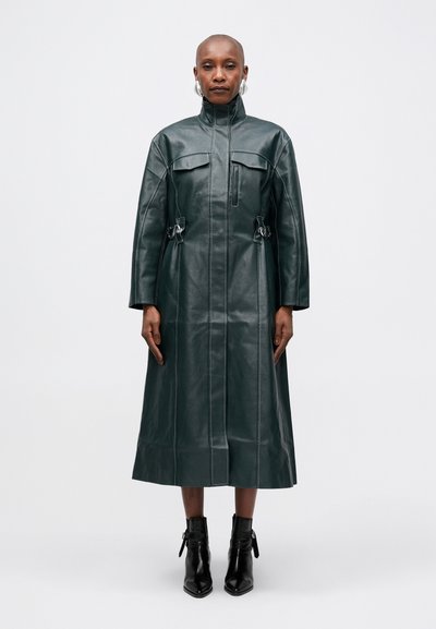 Cappotto lungo verde in pelle con alto colletto, due tasche sul petto e dettagli in vita arricciati. Texture liscia con cuciture visibili. Stivali neri alla caviglia.