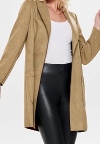 Beige blazer lavet af blødt, struktureret materiale, med to forlommer og et strømlinet, åbent design. Sorte leggings under.