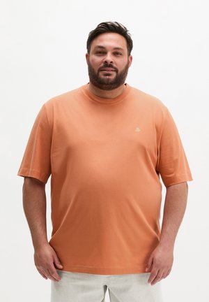 Mann mit kurzen dunklen Haaren und Bart, trägt ein oranges T-Shirt und helle Hosen, steht vor einem schlichten weißen Hintergrund.
