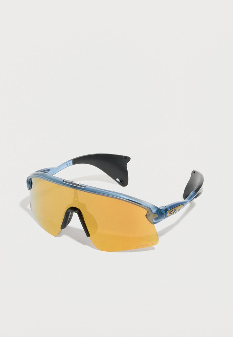 Αθλητικά γυαλιά ηλίου με μπλε πλαίσιο, φακό τύπου wraparound χρώματος κεχριμπαριού και μαύρες λάστιχες στις άκρες των βραχίονων και στα στηρίγματα μύτης, σε λευκό φόντο.