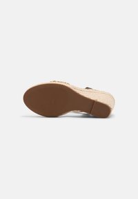 Semelle en caoutchouc marron d'une sandale beige compensée avec un talon tissé style espadrille vue de dessous.