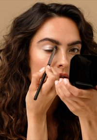 Gros plan sur une personne appliquant un fard à paupières argenté avec un petit pinceau tout en tenant un miroir carré noir, montrant des cheveux noirs ondulés et un teint naturel.