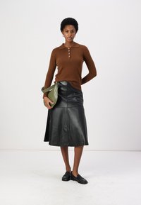 Maglione marrone a maglia con tre bottoni, gonna midi in pelle nera con un design a cucitura e borsa verde. Modella con indosso scarpe nere senza laccio.