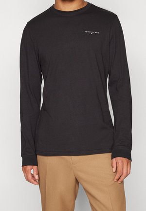 Homme portant un t-shirt noir à manches longues avec col rond et le logo « Tommy Jeans » sur la poitrine, ainsi qu'un pantalon beige, debout devant un fond uni.