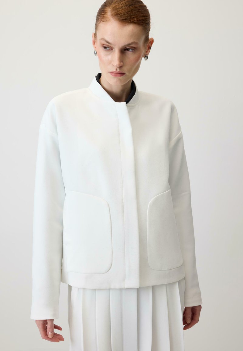 Hvid jakke med høj krave, der har to store forlommer, moderne snit og glat tekstur, kombineret med et plisseret hvidt skørt.