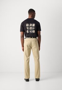 Zwarte t-shirt met een vierkant patroon van bladontwerpen op de achterkant, gecombineerd met beige broek en zwarte schoenen, gezien van de achterkant.