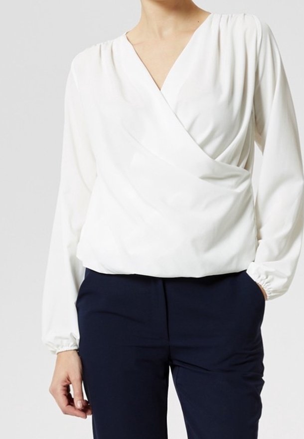 Blouse blanche à manches longues, portefeuille, avec un col en V, des épaules froncées et des poignets élastiques, associée à un pantalon bleu marine. Texture de tissu lisse.