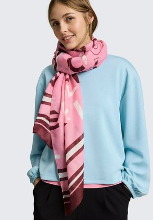 Femme portant un sweat-shirt bleu clair et une grande écharpe rose avec des motifs sombres enroulée autour du cou, les mains dans les poches d'un pantalon noir.