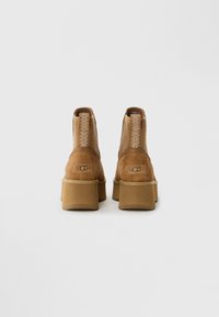 Et par tan semskede UGG platåstøvler med sydde løftestropper, sett fra baksiden på en ensfarget hvit bakgrunn.
