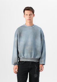 Sweatshirt azul desbotado com gola redonda, mangas longas e bainha desfiada. Usado com jeans pretos desgastados com acentos metálicos.