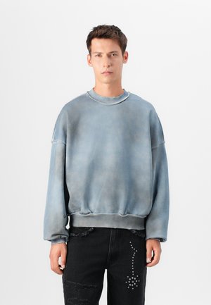 Blå blekt sweatshirt med rund hals, långa ärmar och fransad fåll. Bärs med svarta slitna jeans med metalliska detaljer.