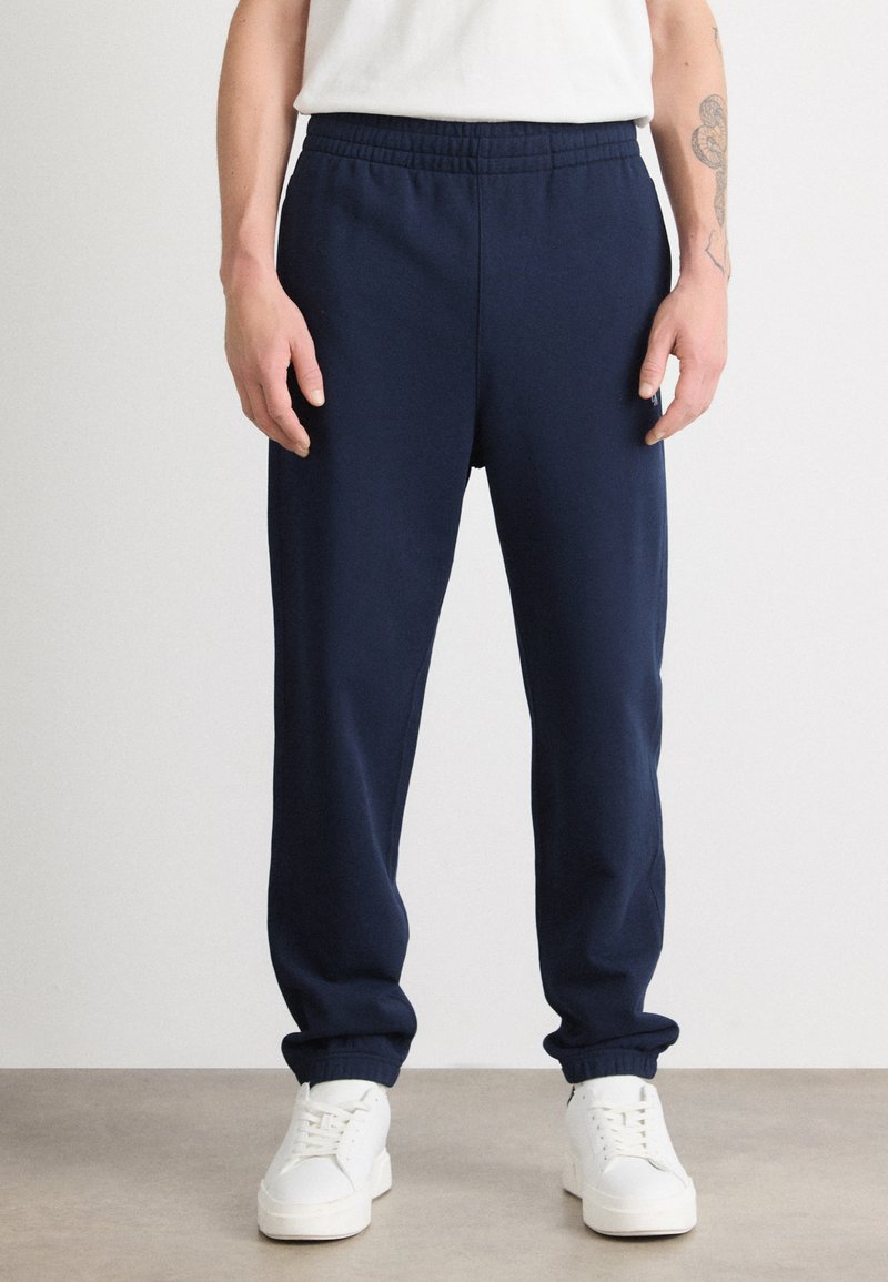 Pantalones de chándal azul marino con cintura elástica, corte ajustado y tobillos ceñidos, combinados con zapatillas blancas sobre un suelo neutro.