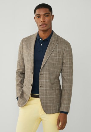 Blazer marrón a cuadros con un corte entallado, llevado sobre una camiseta de polo azul marino, combinado con pantalones amarillos claros y un cinturón negro.