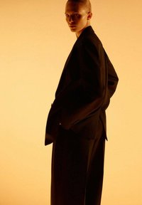 Persoon met slicked-back haar, gekleed in een zwart oversized pak, staand in een driekwart pose tegen een warme gradient achtergrond.