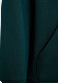 Tissu de sweat-shirt vert foncé avec une texture lisse. Présente une manchette côtelée, une poche latérale et des coutures pour la structure. Design simple et décontracté.