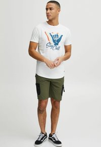 Camiseta blanca de algodón con un colorido gráfico de "Stay Chill" y un signo de mano. Pantalones cortos verde oliva con bolsillos con cremallera. Zapatillas negras.
