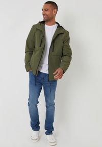 Olivgrüne Jacke mit Kapuze, zwei Fronttaschen und schwarzen Reißverschlüssen, getragen über einem weißen Hemd und blauen Jeans. Weiße Schuhe runden das Outfit ab.