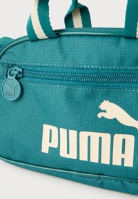Τσάντα αθλητικών ειδών Teal Puma με λευκό λογότυπο Puma, τσέπη με φερμουάρ, και ριγωτές λαβές σε λευκό φόντο.