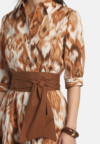 Frau in einem braun-weiß gemusterten knöpfbaren Kleid mit dreiviertel Ärmeln, einem breiten braunen gebundenen Gürtel und einem braunen Armband.