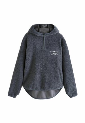 Kvinde stående iført en sort fleece-pullover, sorte leggings, hvide sokker og sølvfarvede sneakers mod en neutral baggrund.