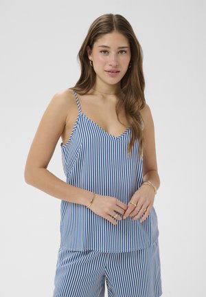 Młoda kobieta z długimi brązowymi włosami, ubrana w niebiesko-biały, paskowany bezrękawnik i pasujące do niego szorty, stojąca ze splecionymi rękami.