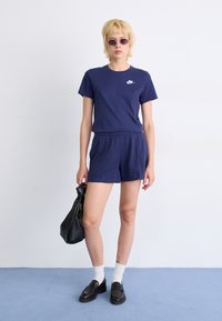 T-shirt en coton bleu marine à manches courtes avec un logo blanc. Associé à un short assorti, des mocassins noirs et un sac noir. Fond clair.