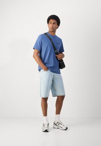 Blauwe crewneck t-shirt, lichte denim shorts en zilveren sneakers. Model draagt een zwarte schoudertas en staat tegen een effen achtergrond.