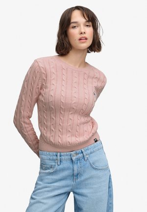 Ung kvinde iført en lyserød kabelstrikket sweater og lyseblå højtaljede jeans, står med hænderne bag ryggen mod en hvid baggrund.