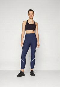 Tengerészkék leggings magas derékkal és fehér díszítésekkel. Fekete sportmelltartóval és fekete sportcipővel párosítva. Simogató, rugalmas anyag.