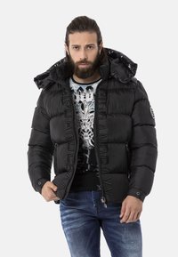 Schwarze Daunenjacke mit großer Kapuze, Reißverschluss vorne, markanter Nahtführung und Logo-Akzent am Ärmel. Trägt man über einem T-Shirt mit Grafikdruck.