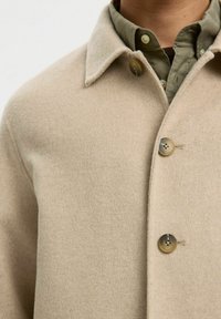 Manteau en mélange de laine beige avec un col pointu et trois grands boutons. En dessous, une chemise boutonnée vert olive est partiellement visible.