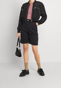 Blouson aviateur noir avec poches zippées, haut rose côtelé, short noir taille haute, chaussures noires à franges, chaussettes dorées et petit sac noir.