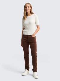 Top corto de manga corta blanco con textura acanalada, combinado con pantalones de pana marrones y zapatillas blancas. El modelo está de pie con las manos en los bolsillos frente a un fondo claro.