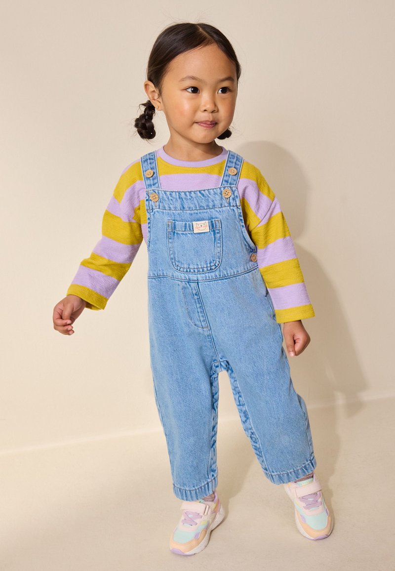 Junges Mädchen mit geflochtenem Haar, das ein gelb-lila gestreiftes Shirt, blaue Jeans-Latzhose und pastellfarbene Turnschuhe trägt, vor beigem Hintergrund steht.