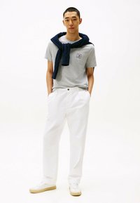 Camiseta gris de manga corta con logo, pantalones anchos blancos y un suéter azul marino anudado al cuello. Zapatillas blancas con suelas beige.