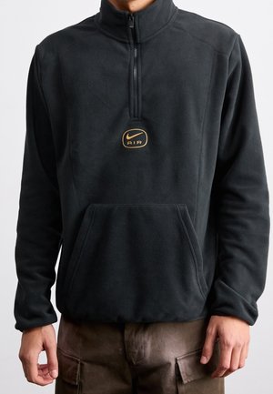 Person trägt schwarzen Halbreißverschluss-Fleece-Sweatshirt mit Fronttasche und gelbem Nike Air-Logo, kombiniert mit braunen Cargohosen.