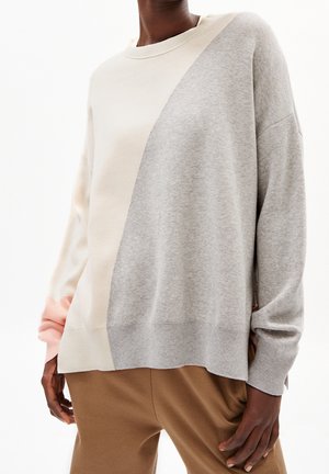 Personne portant un pull ample à blocs de couleurs crème, gris et rose clair, avec un pantalon marron, sur un fond blanc.