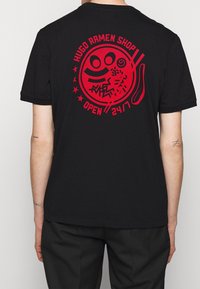 T-shirt en coton noir avec un grand graphique rouge au dos, représentant des images et du texte sur le thème d'un magasin de ramen. Manches courtes, design col rond.