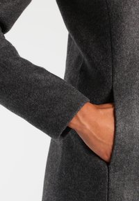 Abrigo de lana gris oscuro con corte ajustado, mangas largas y un bolsillo lateral. La textura parece suave y ligeramente afelpada.