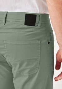 Hellegrüne Hose aus glattem Stoff, mit einem schwarzen Logo-Patch am Bund, zwei Taschen und kontrastierenden Nähdetails.