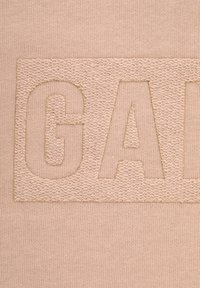Beige bomullstyg med texturerad broderi av ordet "GAP" i en rektangulär lapptäck, med en slät bakgrund och något upphöjda bokstäver.