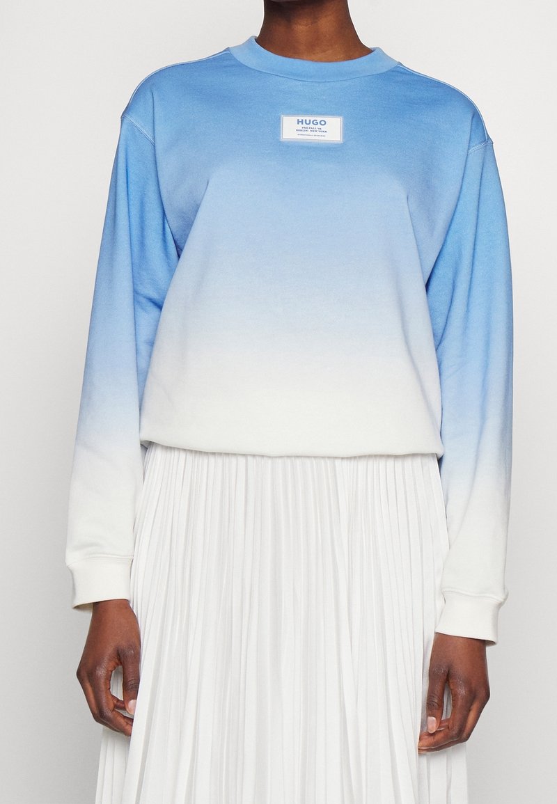 Sudadera azul claro con un diseño ombre que se desvanece hasta el blanco, con mangas largas y un parche de logo en el pecho. Falda blanca plisada visible por debajo.