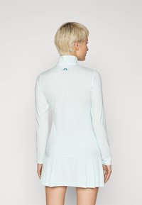 Light blue long-sleeve dress with a high collar and pleated skirt. Features a small logo in teal on the back. Smooth, stretchy fabric.