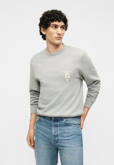 Νέος άνδρας με σγουρά μαλλιά, που φοράει ένα ανοιχτό γκρι φούτερ με λευκό κείμενο λογότυπου και μπλε τζιν, κοιτάζοντας πλάγια, με το χέρι στην τσέπη.