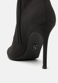 Svart høyhælte støvletter med glatt stoffoverdel, sidelukking med glidelås og spiss tå. Har en slank stilettohæl og merket detalj på sålen.