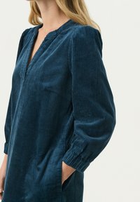 Dunkeltürkisses Cord-Minikleid mit V-Ausschnitt, langen Puffärmeln und Seitentaschen. Das strukturierte Gewebe hat vertikale Rippungen für zusätzliche Details.