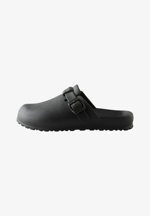 Schwarzer Slip-On-Clog mit strukturierter Sohle und verstellbarem Riemen mit quadratischer Schnalle, seitlich auf weißem Hintergrund dargestellt.