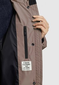 Giacca isolante marrone con interno nero, dotata di una tasca con zip e di un'etichetta che recita "Un abbraccio caldo fatto di stoffa."