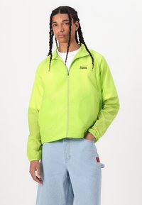 Lightweight, bright green jacket with a zippered front, elastic cuffs, and two circular pockets. Features a logo on the left chest.